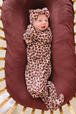 A smiling baby wears an Antique Floral Ribbed Bamboo Knot Gown, featuring a floral pattern and matching headband, nestled in a cozy, brown bamboo bassinet.
