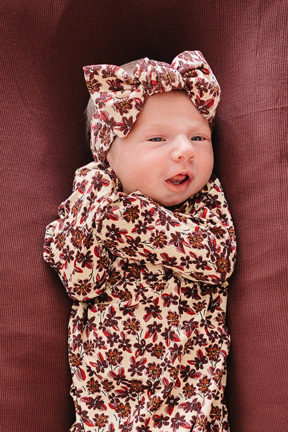 A newborn baby dressed in a floral-patterned bamboo outfit lies on a brown ribbed cushion inside a wicker bassinet, showcasing soft, gentle fabric perfect for delicate skin.