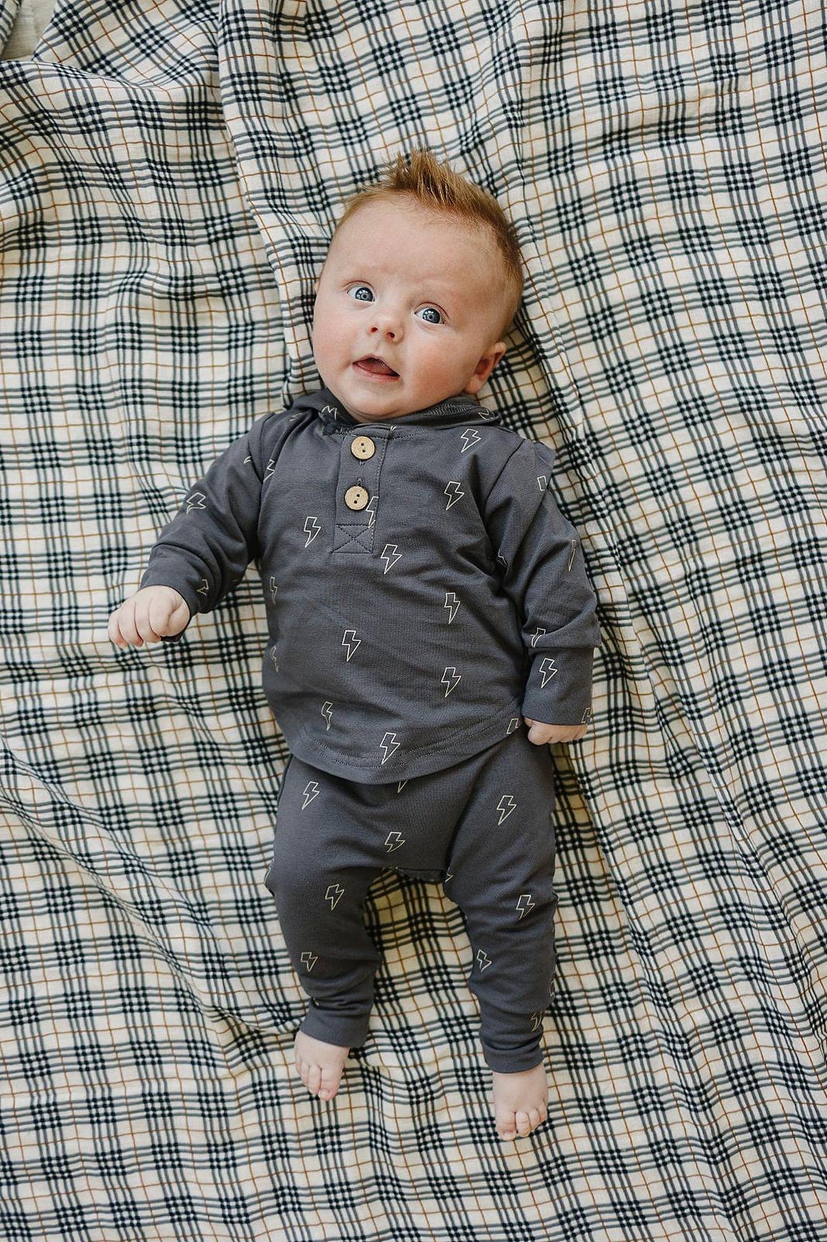 A baby in a navy lightning bolt hooded set lies on a plaid blanket, looking up.