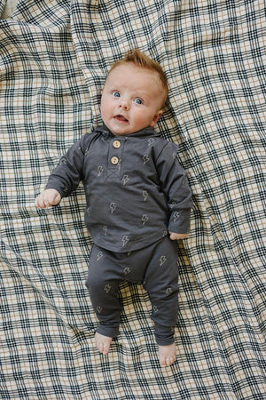A baby in a navy lightning bolt hooded set lies on a plaid blanket, looking up.