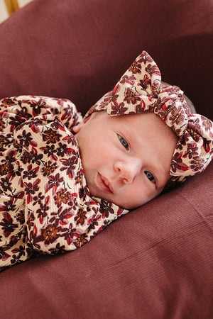 A newborn baby in a floral-patterned ribbed bamboo gown rests on a soft, maroon fabric. The outfit features a matching headband, showcasing a gentle and cozy style for delicate skin.