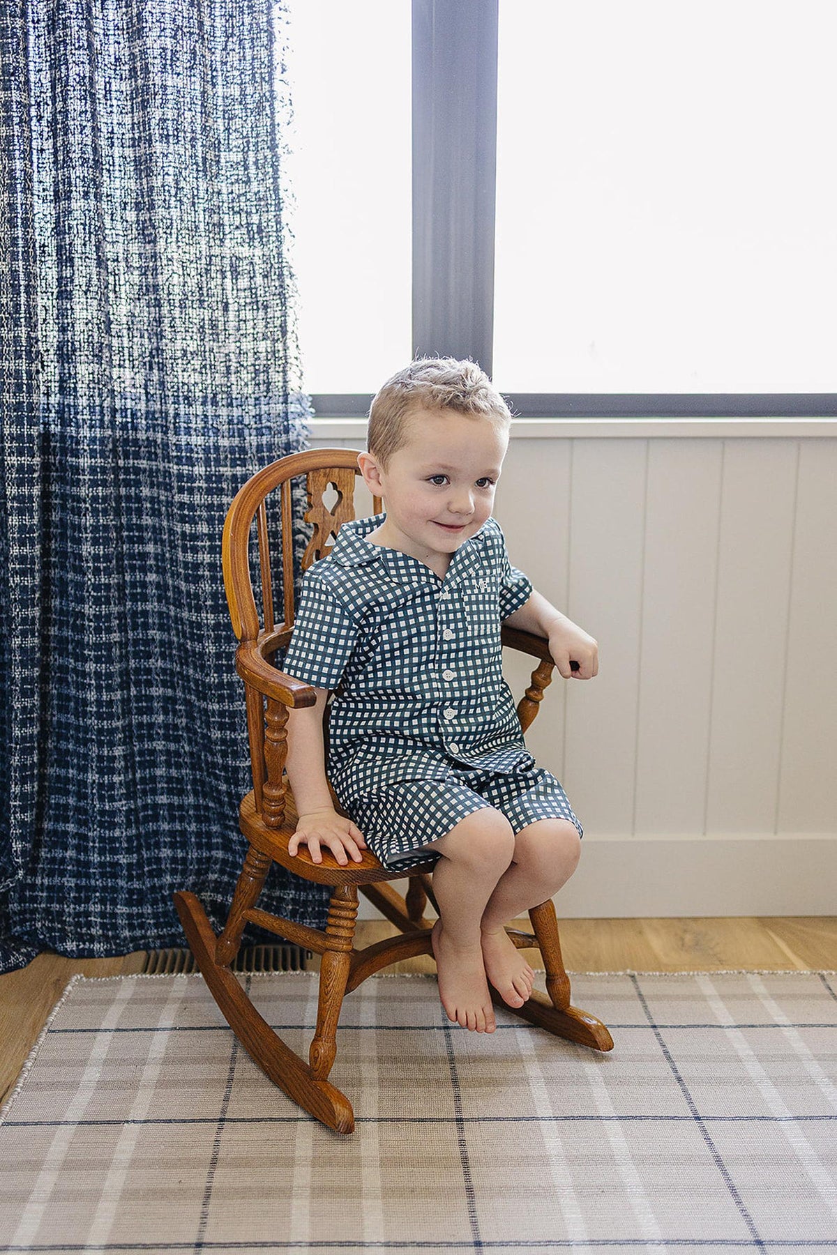 A child in navy gingham pajamas sits on a wooden rocking chair, with a blue curtain in the background.