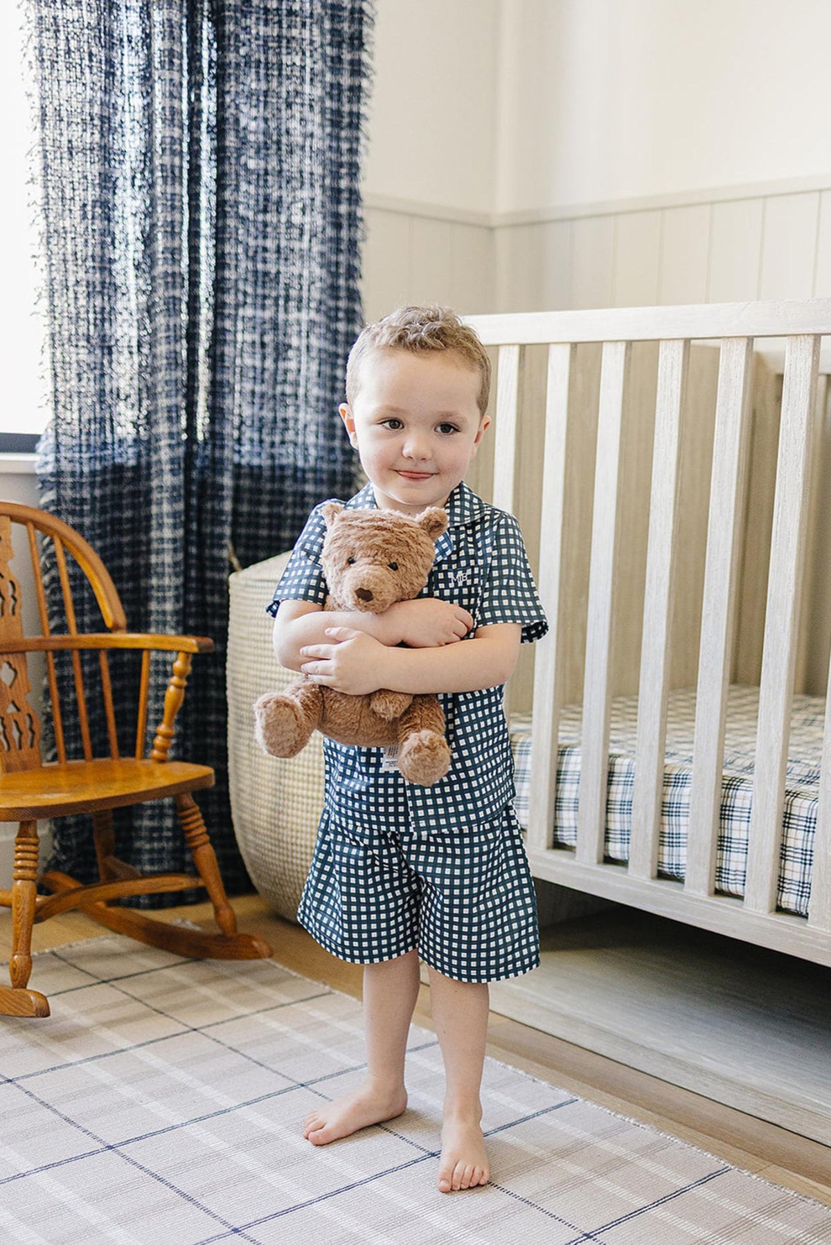 A child in navy gingham pajamas holds a teddy bear, standing in a cozy room with a crib.