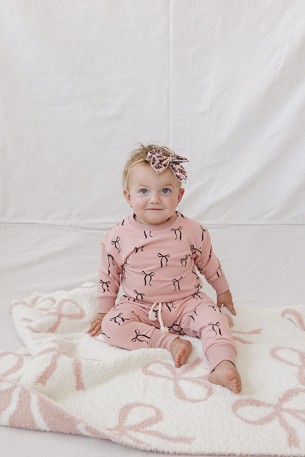A toddler sits on a soft blanket, wearing a blush pink French terry cotton set adorned with a charming bow print.