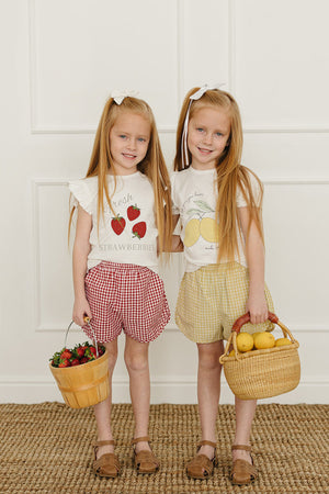 Two young girls stand side by side, one wearing red gingham shorts and the other in yellow gingham shorts, both.