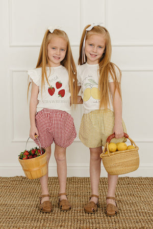 Two young girls stand side by side, wearing playful graphic tees featuring strawberries and lemons, paired with checkered shorts. They hold baskets filled with strawberries and lemons, smiling cheerfully.