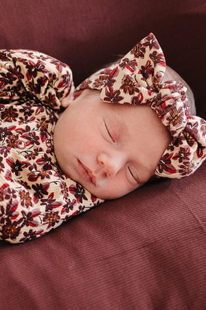 A peacefully sleeping newborn wears a floral-patterned outfit and matching head wrap, both featuring soft, breathable fabric. The setting has a warm, cozy atmosphere with rich maroon bedding.