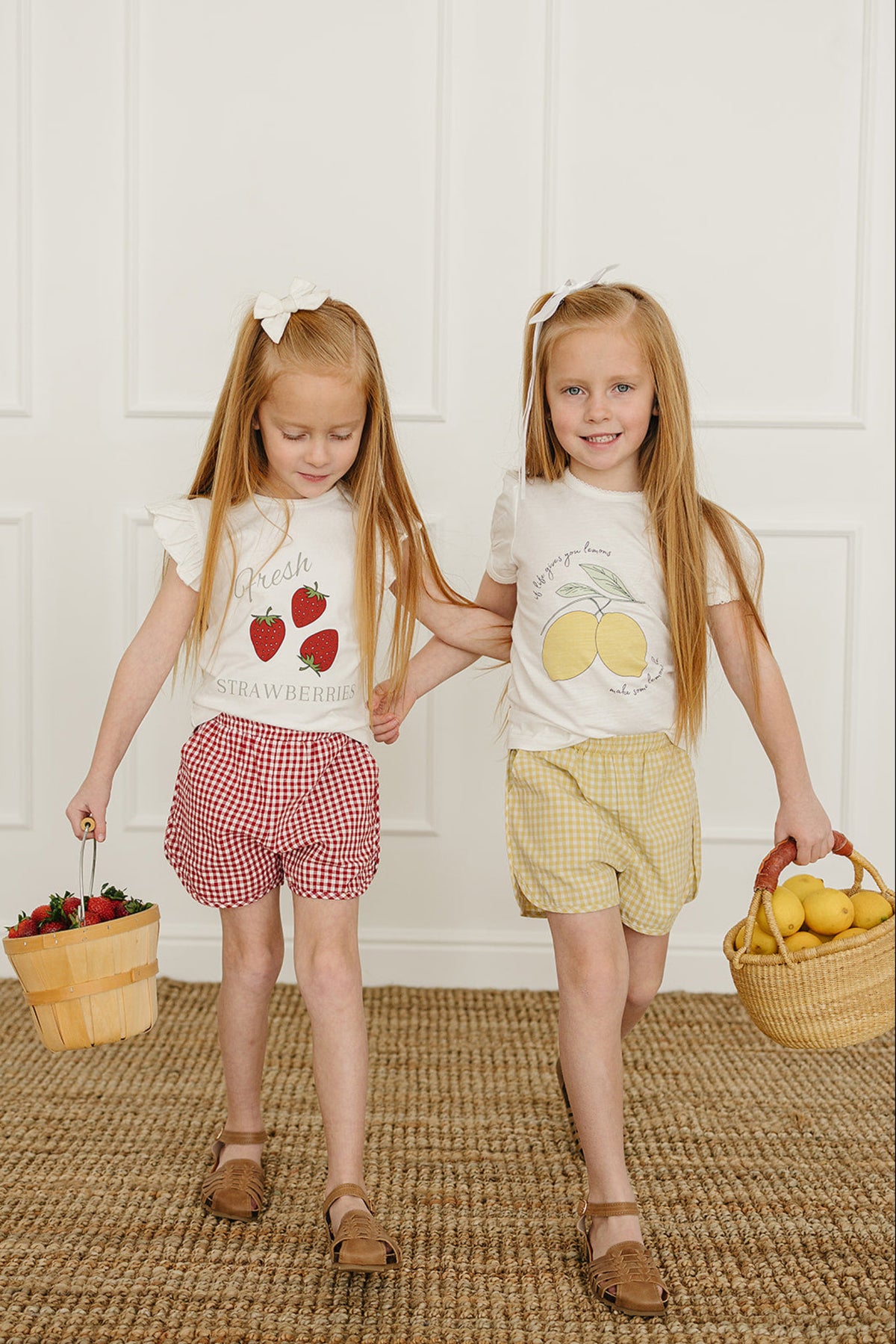 Two young girls are walking together, one wearing a white strawberry graphic tee and the other in a lemon graphic tee, both paired with cheerful, gingham shorts.