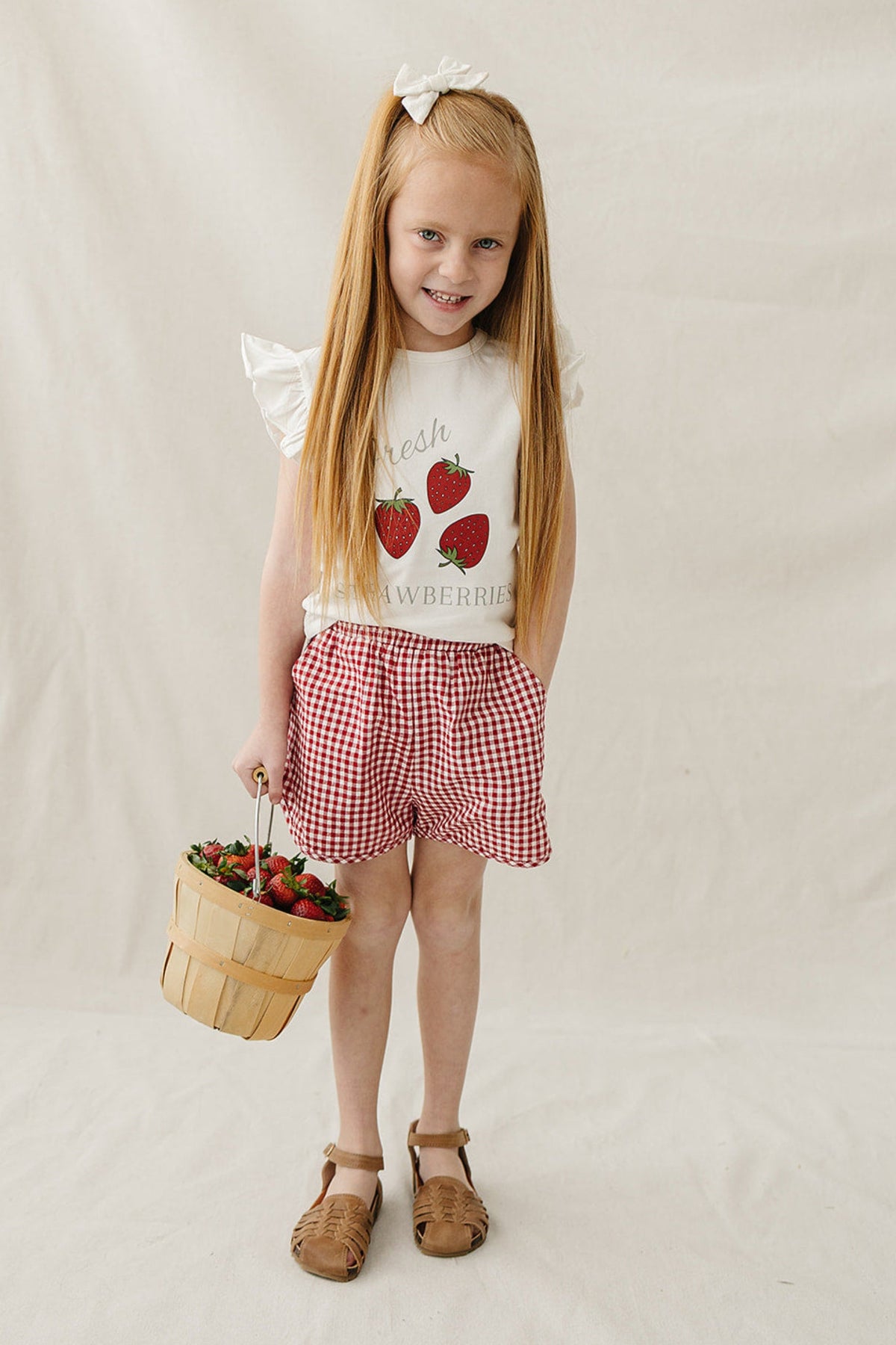 A young girl with long, straight hair wears a white ruffle-sleeve t-shirt featuring a strawberry graphic and red gingham shorts, holding a basket of strawberries against a neutral backdrop.