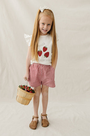 A young girl with long, straight hair wears a white ruffle-sleeve t-shirt featuring a strawberry graphic and red gingham shorts, holding a basket of strawberries against a neutral backdrop.