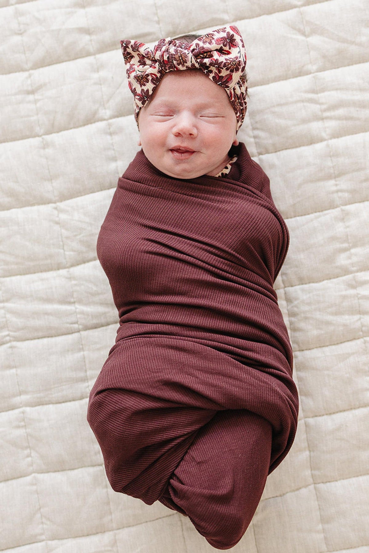 A newborn is swaddled in a deep burgundy ribbed bamboo wrap, featuring a matching floral head wrap.