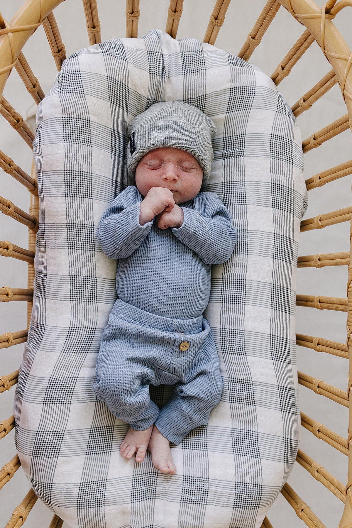 A peacefully sleeping newborn swaddled in a dusty blue waffle cotton set, featuring a long sleeve bodysuit and relaxed pants, rests in a rattan bassinet lined with a checkered blanket.