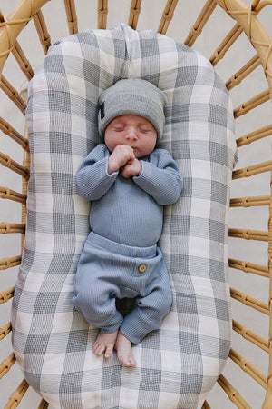 A peacefully sleeping newborn swaddled in a dusty blue waffle cotton set, featuring a long sleeve bodysuit and relaxed pants, rests in a rattan bassinet lined with a checkered blanket.