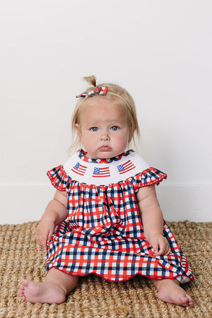 A baby girl sits on a textured, natural fiber rug, wearing a red and white gingham dress with an embroidered American flag collar.