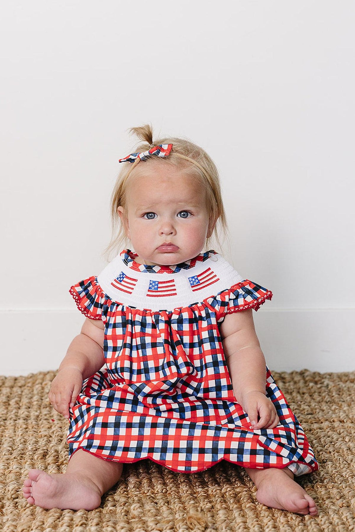 A baby sits on a textured, natural fiber rug, wearing a red and white gingham dress with a decorative collar.