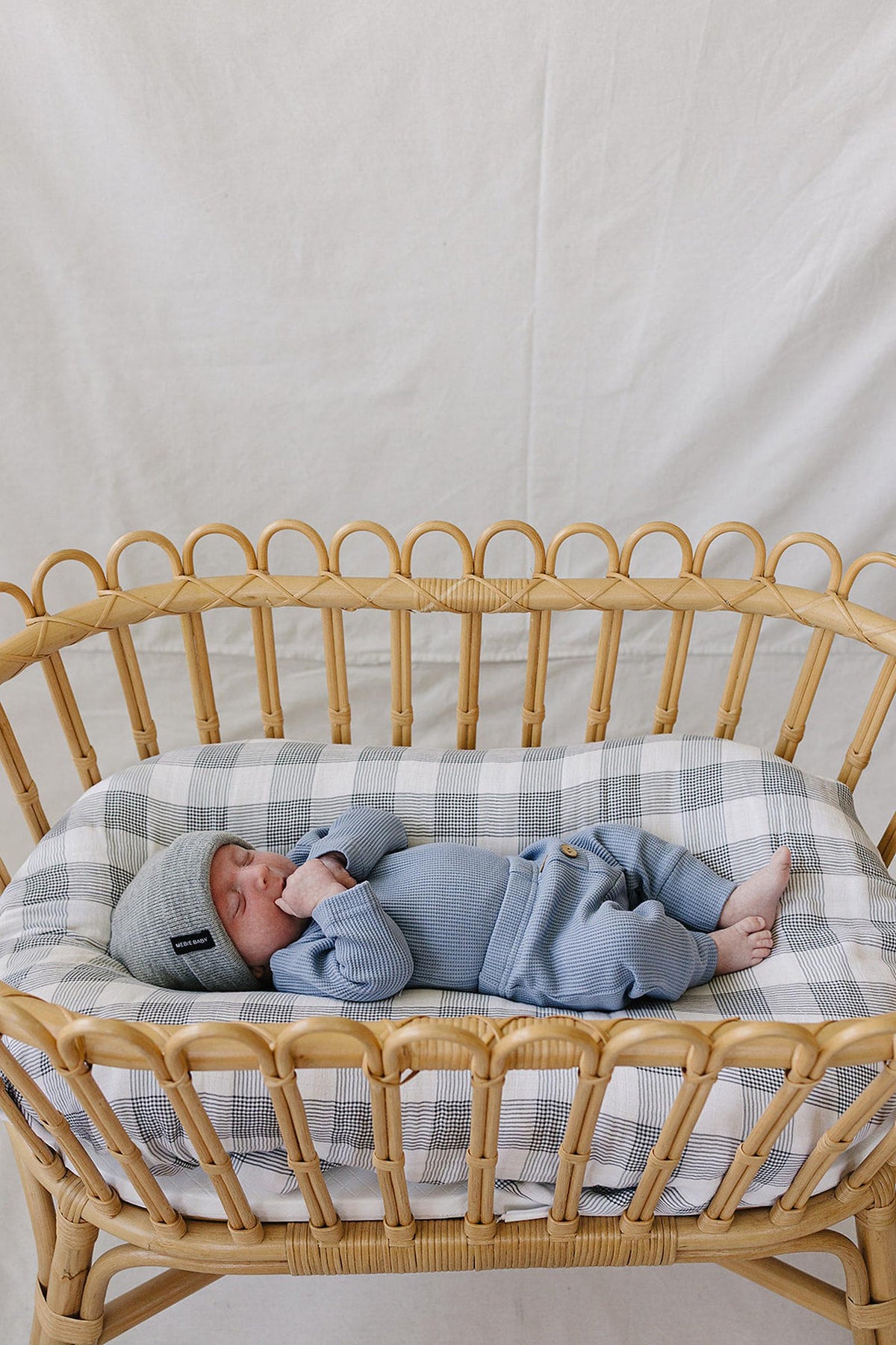 A peaceful baby lies on a checkered blanket in a rattan bassinet, wearing a soft dusty blue waffle cotton outfit and a gray hat, showcasing a cozy and stylish look.