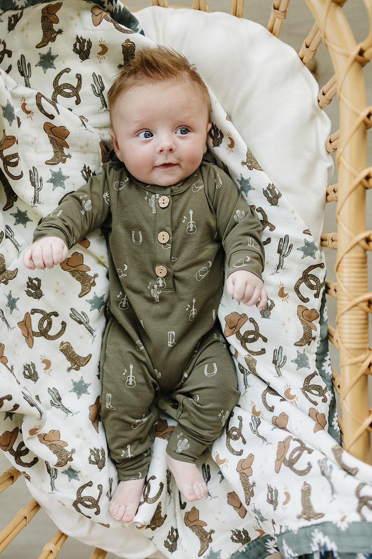 Baby wearing an olive green romper with western patterns, lying on a themed blanket in a wicker basket.