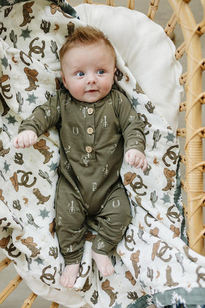 Baby wearing an olive green romper with western patterns, lying on a themed blanket in a wicker basket.