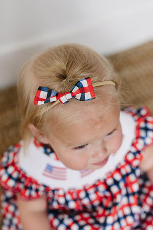 A young child wears a red, white, and blue plaid hair bow made of fabric, complementing her ruffled dress adorned with American flags. The setting features a natural woven rug.