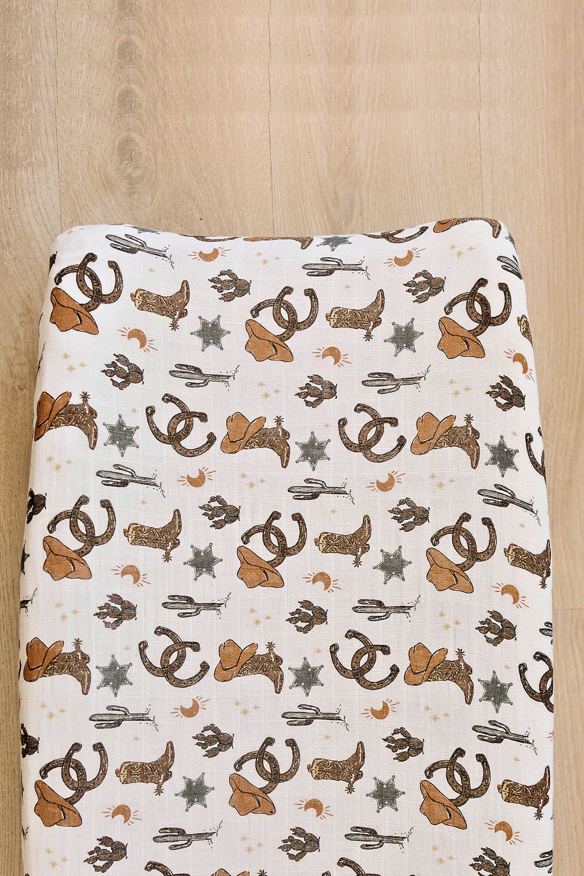 A muslin changing pad cover featuring a playful cowboy theme, showcasing brown hats, stars, and horseshoes on a light background. Made from 100% cotton, its soft and breathable for babies.