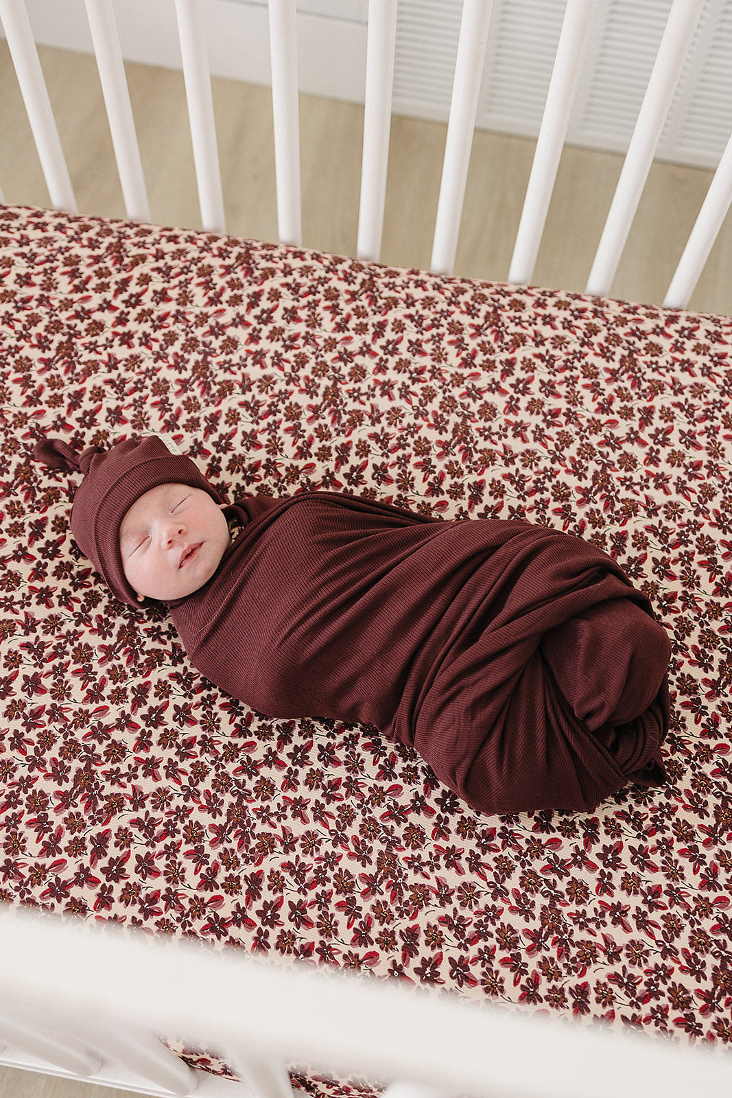 A peacefully sleeping baby is swaddled in a deep burgundy fabric on a floral-patterned crib sheet, showcasing a soft, cozy environment for rest. The crib features a clean, modern design.