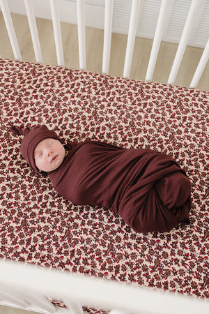 A peacefully sleeping baby is swaddled in a deep burgundy fabric on a floral-patterned crib sheet, showcasing a soft, cozy environment for rest. The crib features a clean, modern design.