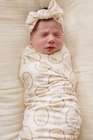 A peacefully sleeping newborn is wrapped in a soft, beige swaddle featuring a vintage bow print. The swaddle is complemented by a matching headband, both made from bamboo fabric.