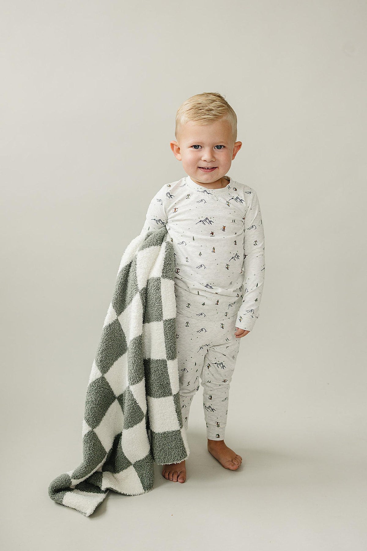 A smiling toddler wears a ski print pajama set and holds a green and white checkered blanket.