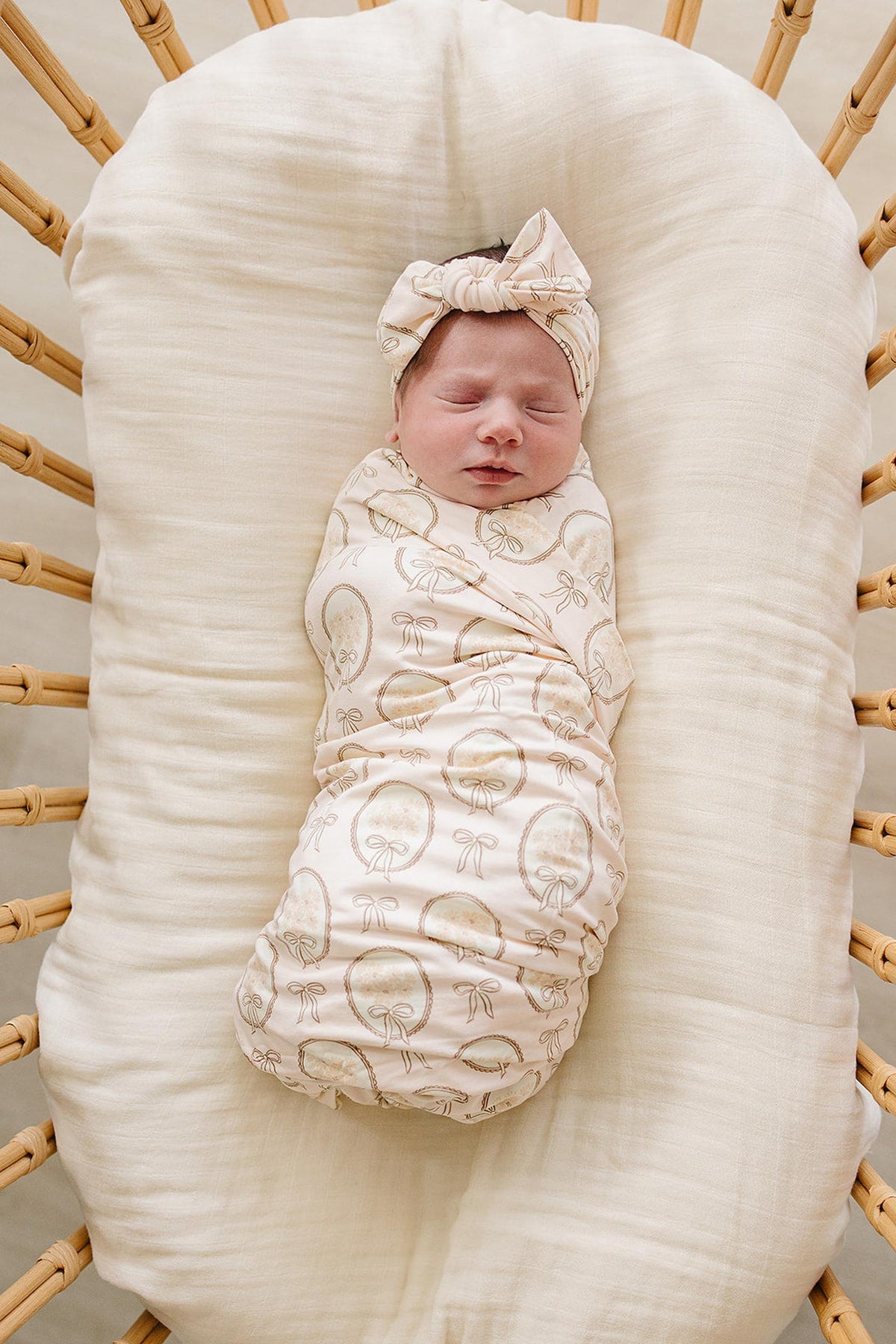 A peacefully sleeping baby is swaddled in a light-colored, patterned bamboo stretch swaddle. The swaddle features a matching headband, complementing the cozy, natural setting of a woven bassinet.