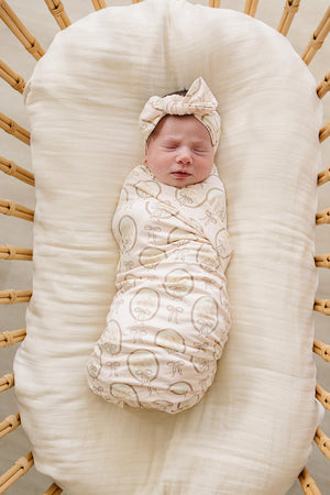 A peacefully sleeping baby is swaddled in a light-colored, patterned bamboo stretch swaddle. The swaddle features a matching headband, complementing the cozy, natural setting of a woven bassinet.
