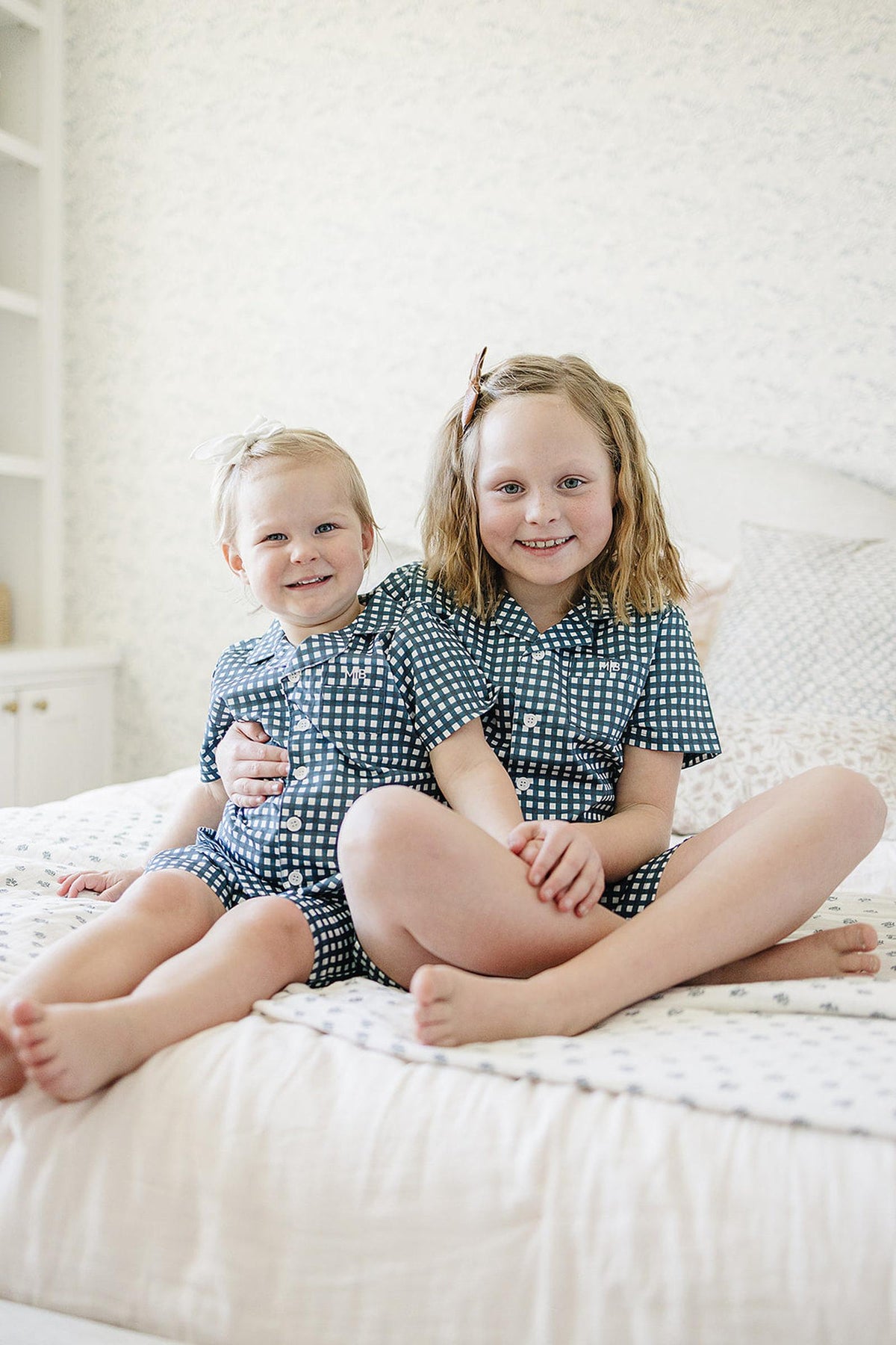 Two young children sit on a bed wearing matching navy gingham pajama sets, smiling in a cozy bedroom.