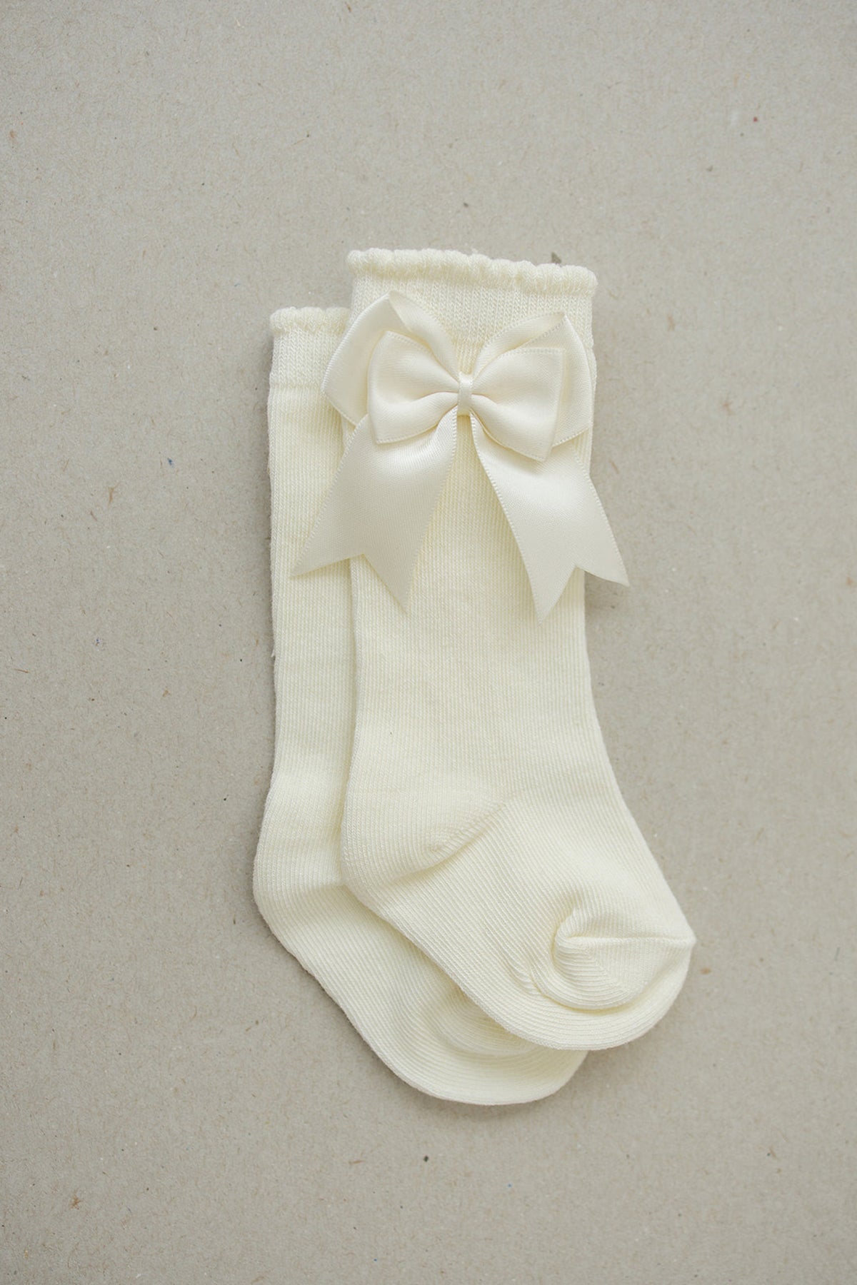 Cream-colored baby socks with a large satin bow, made from soft cotton, displayed on a neutral background.