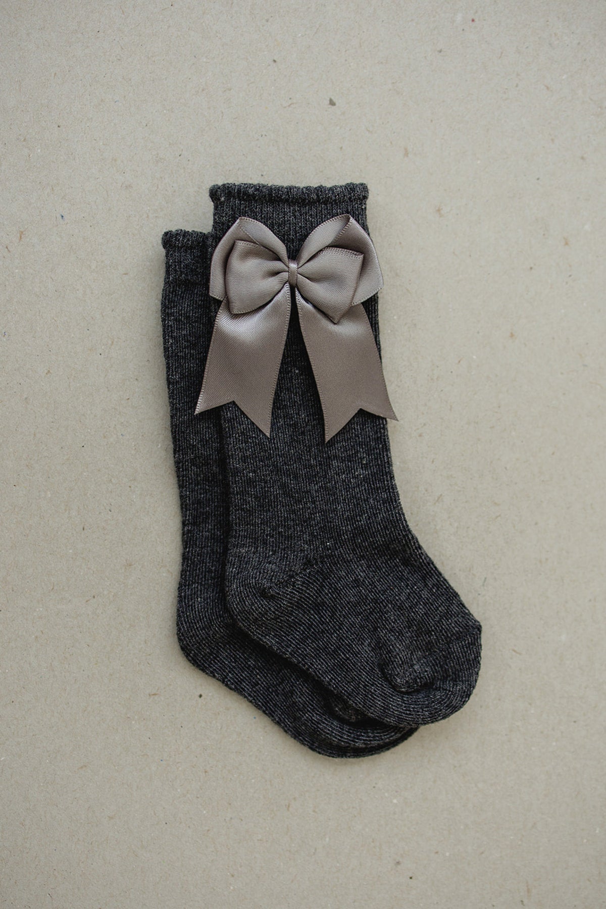 Dark gray high-rise socks with a large, taupe satin bow on a neutral background, designed for toddlers.