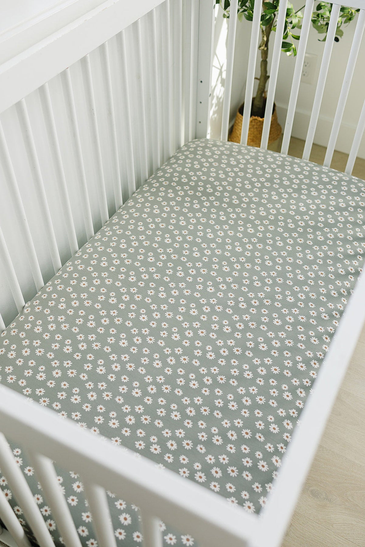 Light green crib sheet with a white daisy pattern, placed in a white wooden crib against a light-colored floor.