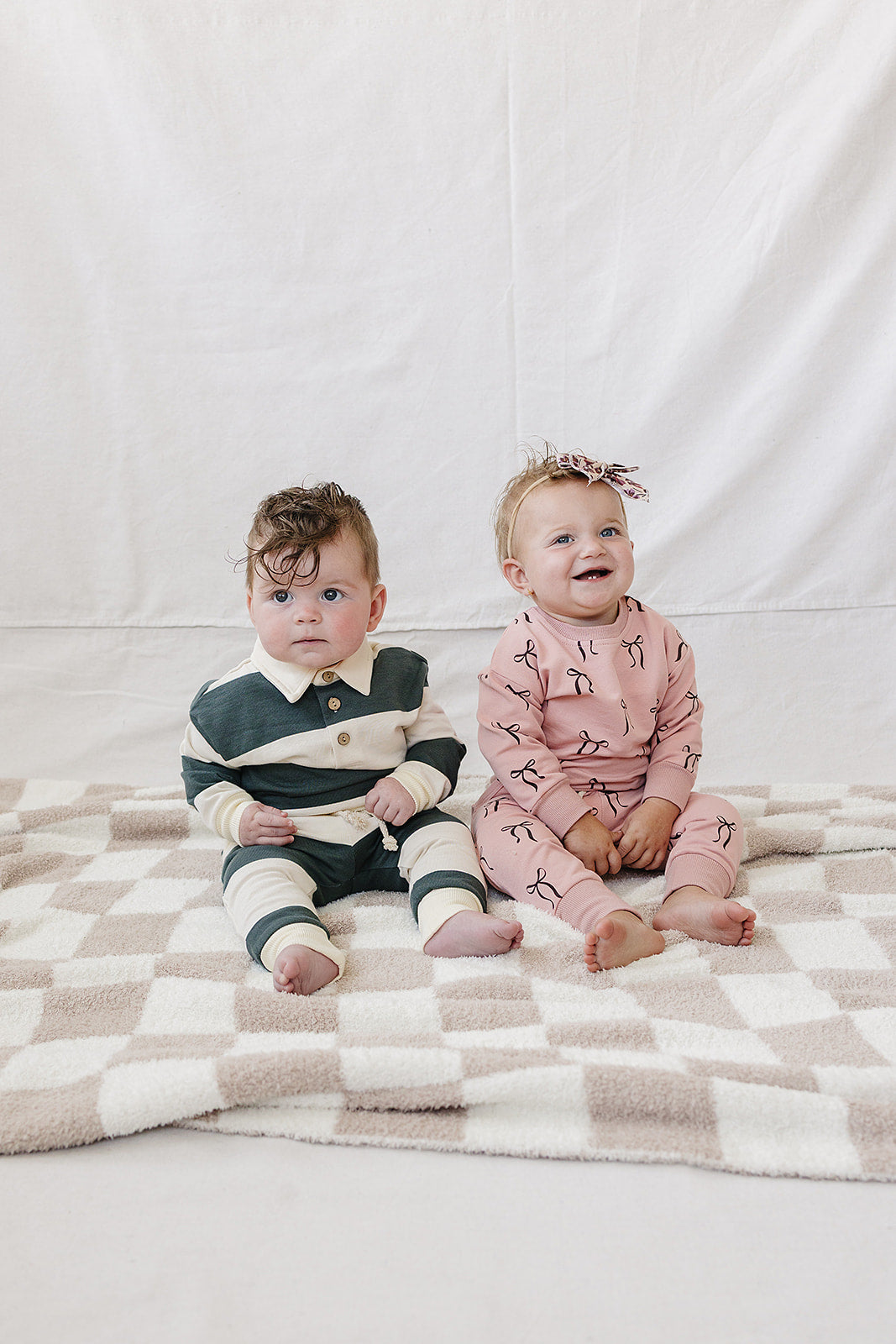 Two babies sit on a soft, checkered blanket, one wearing a green and white striped outfit and the other in a blush pink set featuring bow prints.