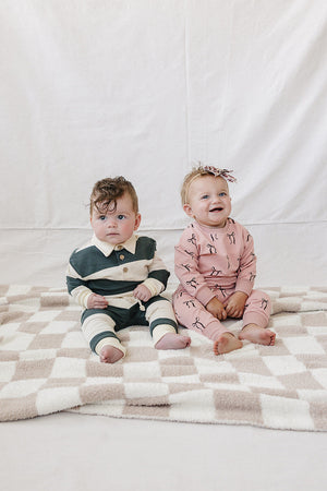 Two babies sit on a soft, checkered blanket, one wearing a green and white striped outfit and the other in a blush pink set featuring bow prints.