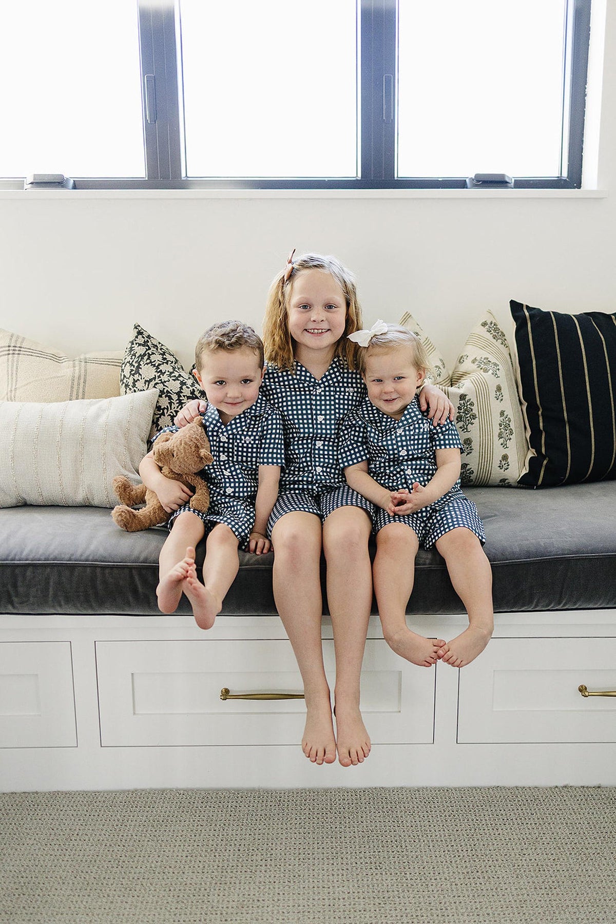 Three children in navy gingham pajamas sit on a cushioned bench, with one holding a teddy bear.