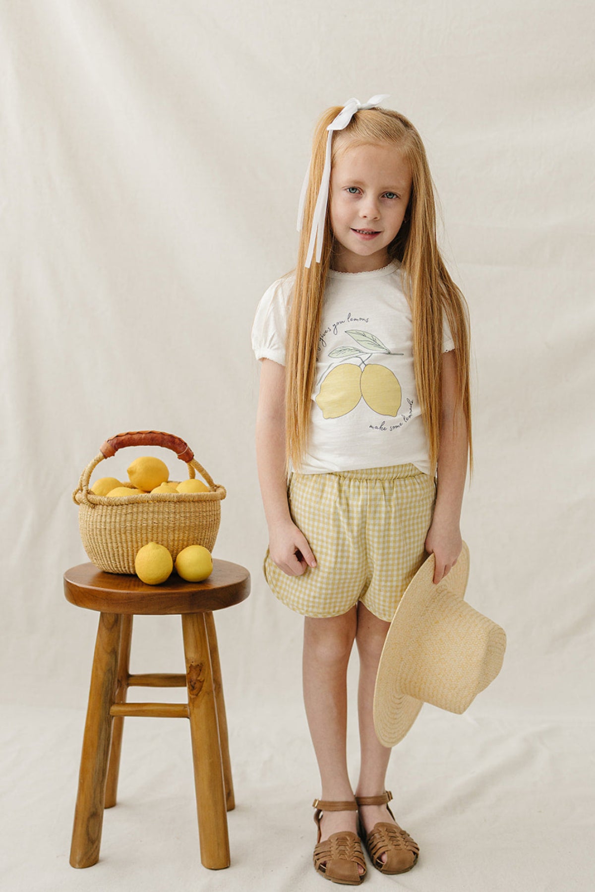 A girl in yellow gingham shorts and a lemon-themed shirt stands beside a wicker basket filled with lemons.