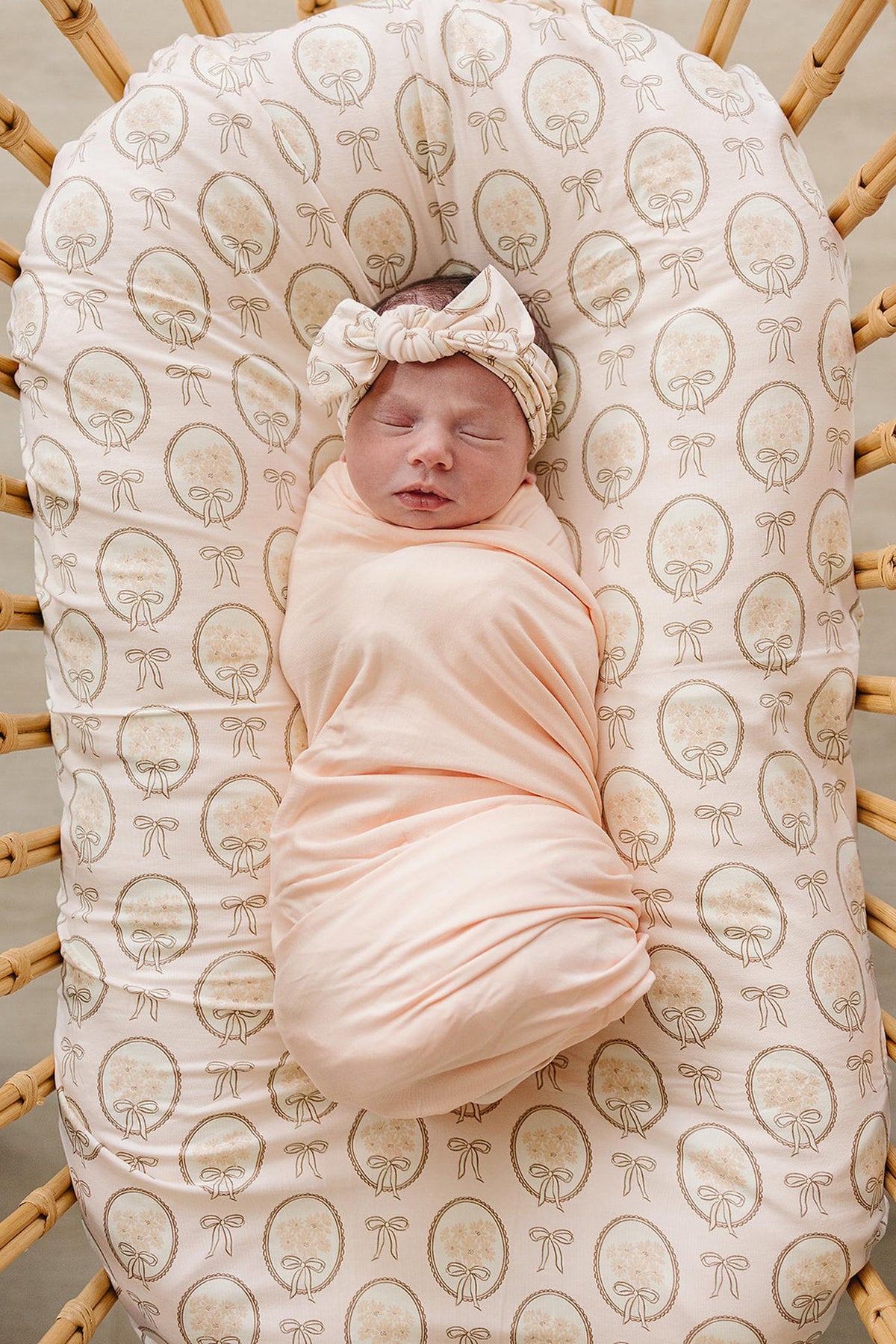 A peaceful newborn is swaddled in a soft, peach-colored fabric, wearing a matching head wrap, resting on a patterned baby lounger within a woven bassinet.