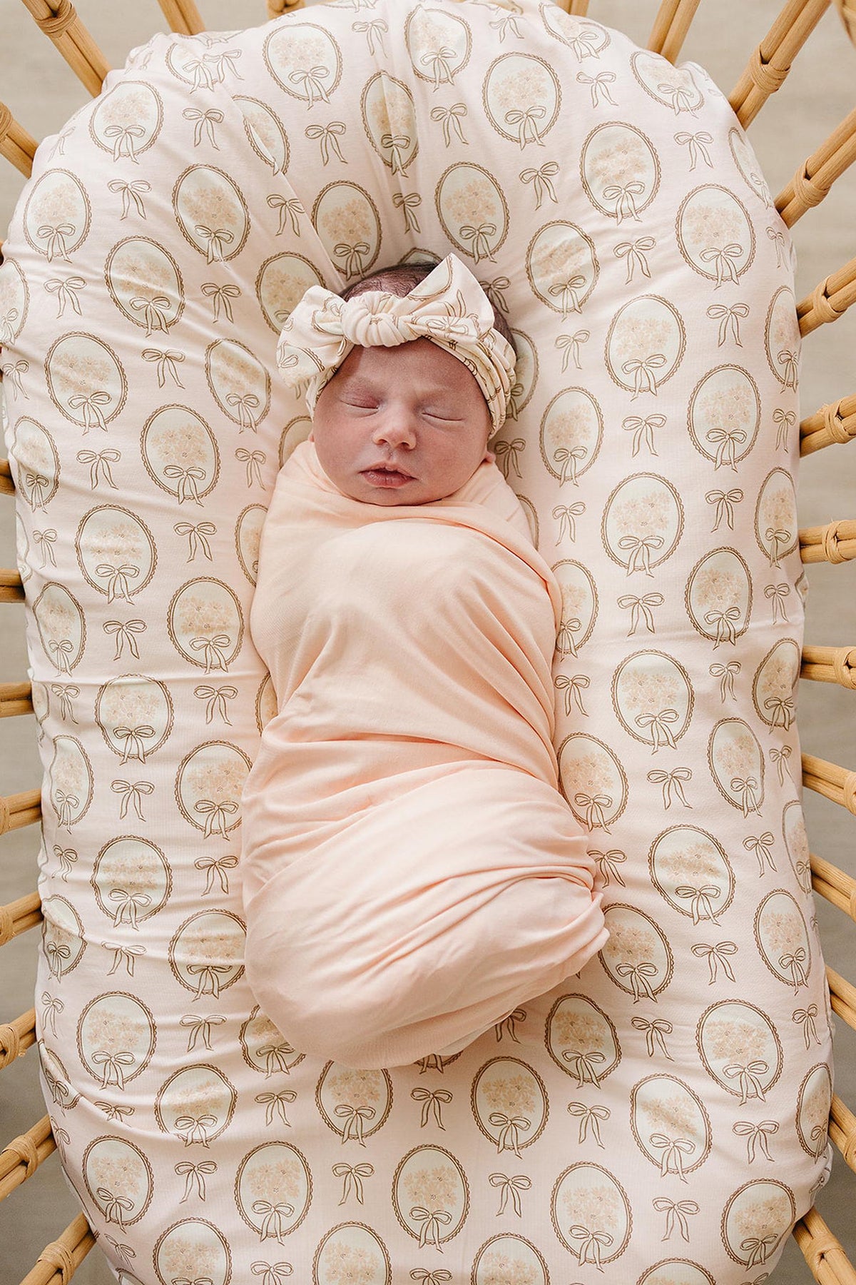 A peaceful newborn is swaddled in a soft peach bamboo blanket, lying in a woven bassinet with a delicate printed liner.
