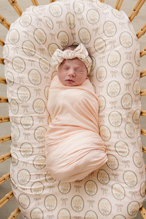 A peaceful newborn swaddled in a soft peach-colored fabric lies in a woven bassinet. The swaddle features a delicate vintage bouquet print, adding a charming touch to the cozy setting.