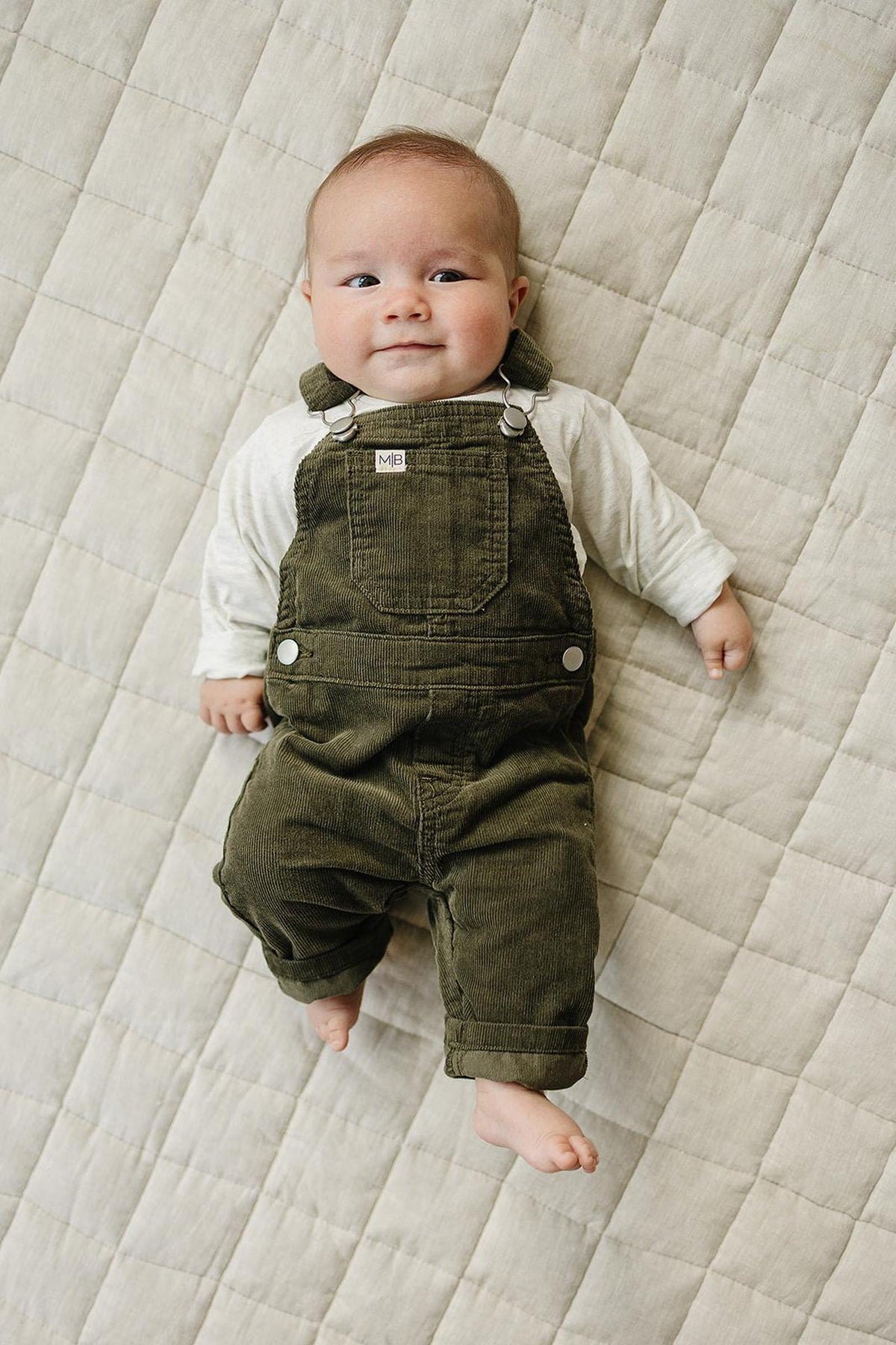 Baby wearing green corduroy overalls lies on a quilted beige blanket, showcasing a cute and cozy outfit.
