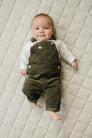 Baby wearing green corduroy overalls lies on a quilted beige blanket, showcasing a cute and cozy outfit.