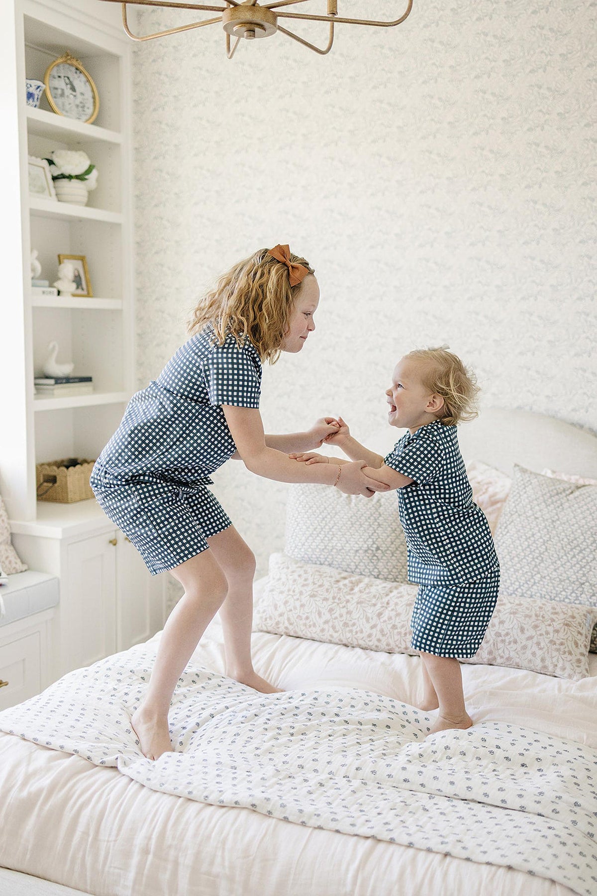 Two children wearing navy gingham pajamas joyfully play on a bed with white patterned bedding in a cozy room.