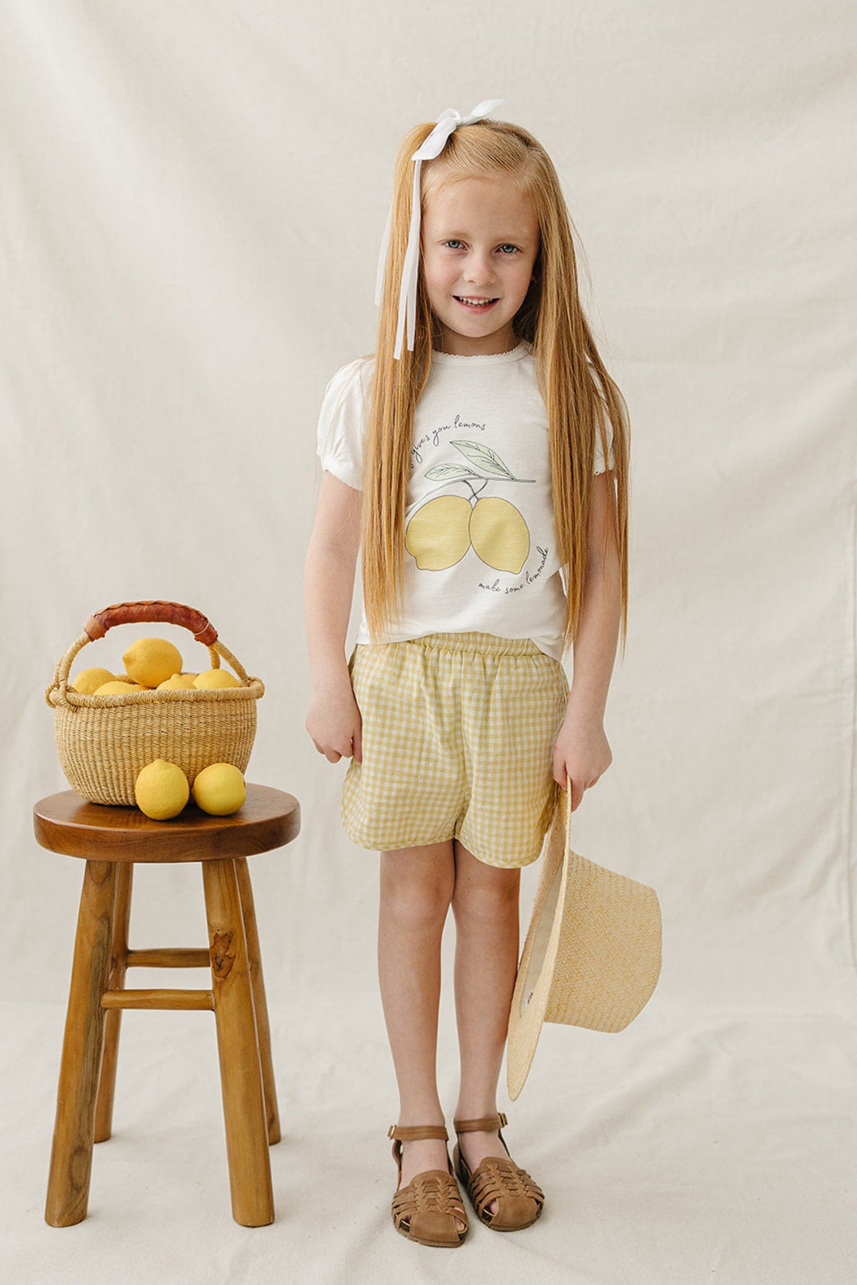A young girl stands on a neutral backdrop, wearing a white T-shirt with a lemon design and yellow checkered shorts.