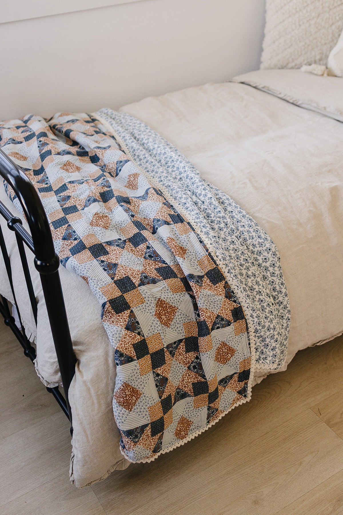 A patchwork quilt with blue, brown, and floral patterns drapes over a beige bedspread on a metal bed frame.