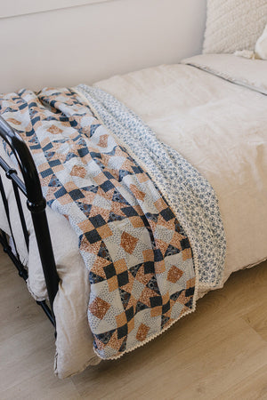 A patchwork quilt with blue, brown, and floral patterns drapes over a beige bedspread on a metal bed frame.