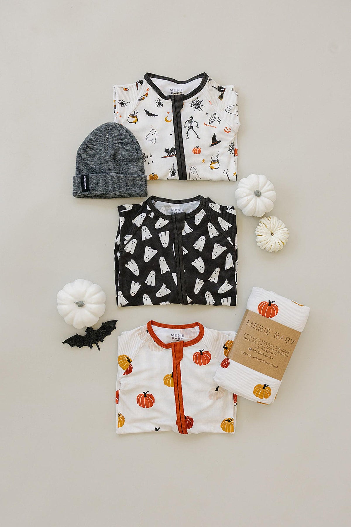 Three Mebie Baby pajamas with Halloween patterns, a gray beanie, and decorative pumpkins on a neutral background.