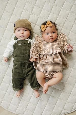 Two babies lying on a quilted blanket; one wears green corduroy overalls and a beanie, the other in a gingham.