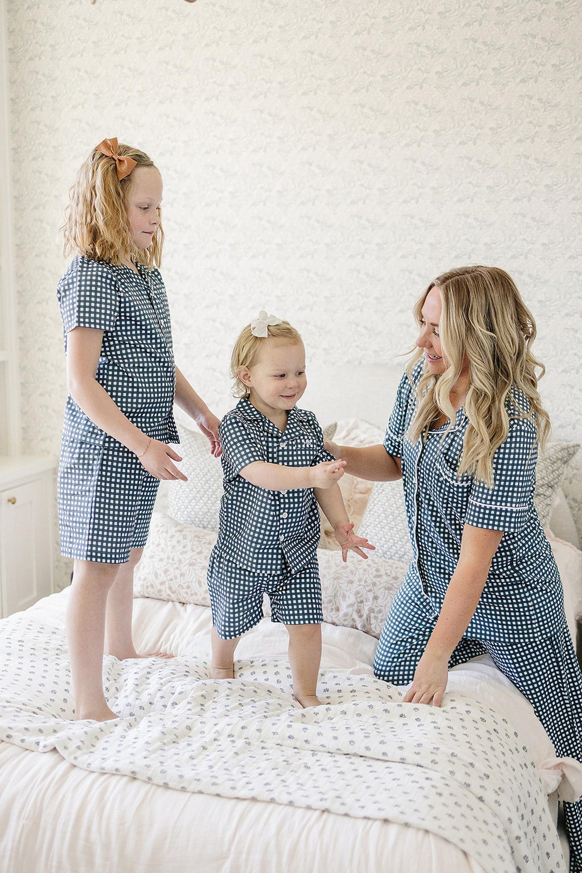 A woman and two children in navy gingham pajamas play on a bed, smiling joyfully in a bright room.